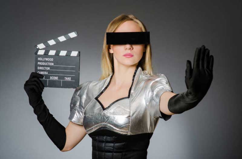 A woman with black gloves and a black rectangle across her eyes, holding a clapperboard in her right hand and gesturing to ‘stop’ with her left hand